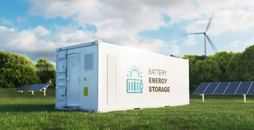 A long cabin with the text "BATTERY ENERGY STORAGE" sits in a lush green field flanked by PV solar panels with a wind turbine turning in the distance